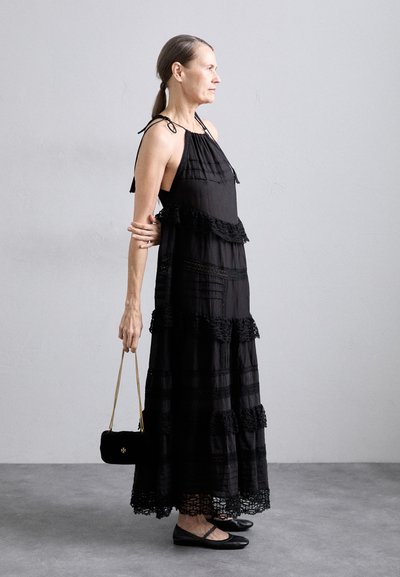 Abito maxi nero con strati di volant e finitura in pizzo, caratterizzato da legami sulle spalle. Abbinato a una piccola borsa nera e a ballerine.