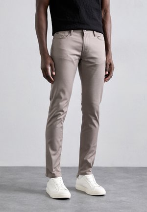 Pantalons beige slim-fit en tissu lisse, avec une taille standard et un design à cinq poches, associés à des baskets blanches.