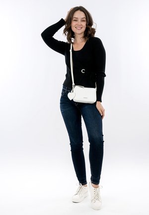 Young woman smiling, wearing black sweater and dark jeans, holding white crossbody bag with heart charm, standing on white background.