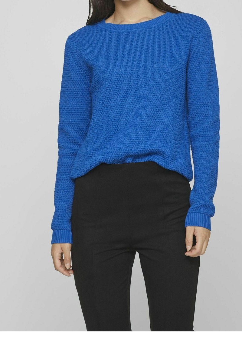 Blå stickad tröja med en texturerad design, rund halsringning och ribbad mudd, ihop med svarta åtsittande byxor.