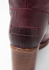 Burgunderfärgade läderankelstövlar med en texturerad yta, som har en brun trähæl och präglad logotyp bak.