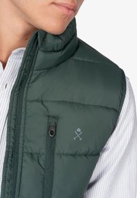 Chaleco inflable verde con cuello alto, bolsillo delantero con cremallera y detalle de logo. Presenta textura acolchada y tela suave.