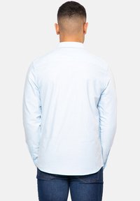Camisa de manga larga azul claro con una yoke trasera, puños abotonados y dobladillo curvado, hecha de una tela suave. Se lleva con jeans oscuros.