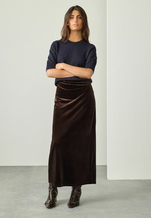 Donna in piedi con le braccia incrociate, indossa un maglione corto blu navy, una maxi gonna in velluto marrone scuro e stivali a punta marrone scuro.