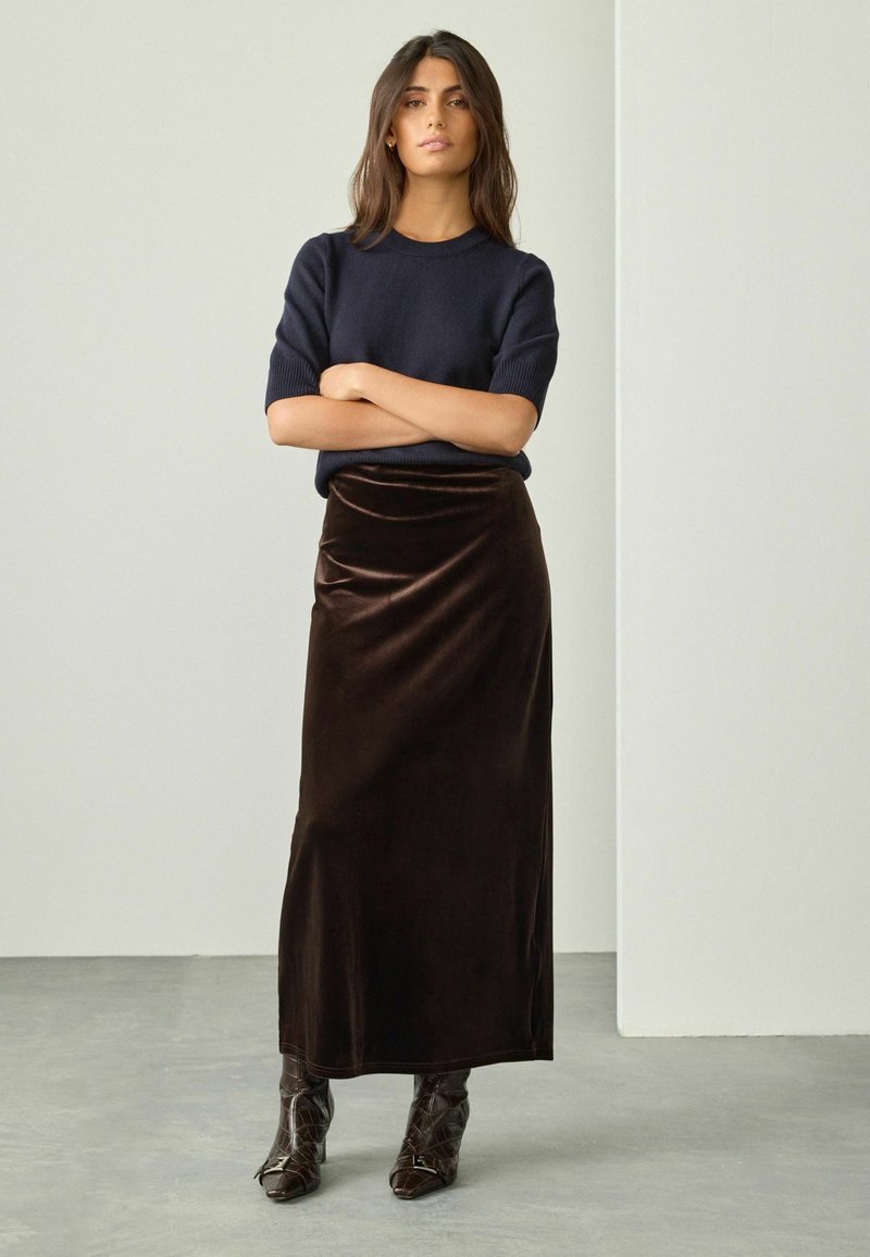 Donna in piedi con le braccia incrociate, indossa un maglione corto blu navy, una maxi gonna in velluto marrone scuro e stivali a punta marrone scuro.