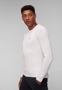Maglione bianco a coste con maniche lunghe, scollatura rotonda e discreto dettaglio del logo. Indossato con pantaloni scuri, caratterizzati da una vestibilità aderente.