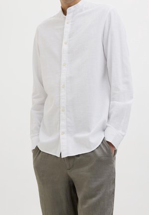 Man draagt wit overhemd met lange mouwen, mandaarin kraag en grijze broek, handen in de zakken, tegen een effen witte achtergrond.