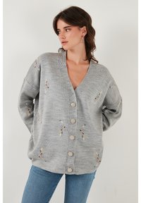 Cárdigan gris con cuello en V, que presenta patrones florales bordados en azul, amarillo y rojo. Seis botones grandes en la parte delantera. Textura suave.