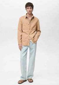 Camicia beige con bottoni, dotata di due tasche sul petto, abbinata a pantaloni larghi azzurri chiari e sandali marroni. Tessuto liscio, vestibilità casual.