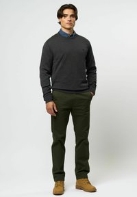 Dunkelgrauer Pullover über einem blauen Hemd mit Kragen, kombiniert mit olivgrünen Hosen und beigen Stiefeln. Das Model steht mit den Händen in den Taschen vor einem neutralen Hintergrund.