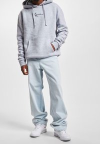 Sudadera gris con capucha, bolsillo delantero y logo negro bordado, combinada con vaqueros holgados azul claro y zapatillas blancas.