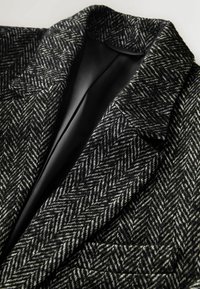 Houndstooth raksta bleizeris melnbaltā vilnā, ar iegrieztu atlokumu un vienu kabatu ar gludu melnu odere.