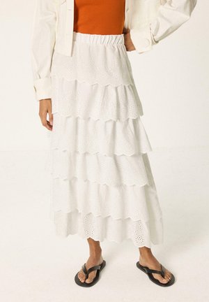 Jupe midi blanche à volants avec broderie anglaise, taille élastique, portée avec un top orange, une veste blanche et des tongs noires.