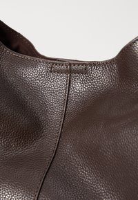 Hnědá kožená taška s texturovaným povrchem, s viditelným prošitím a zaobleným horním okrajem. Jednoduchý design bez kování nebo výrazných doplňků.