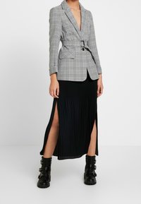 Grå rutigt blazer med djup V-ringning, bälte i midjan och två fickor, kombinerat med en knälång svart kjol med höga slitsar och ankboots.