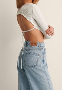 Ein weißes Langarmoberteil mit gerafftem Rücken und Bändern, kombiniert mit hellblauen, hoch taillierten Jeans aus Denim mit einem Lederetikett.