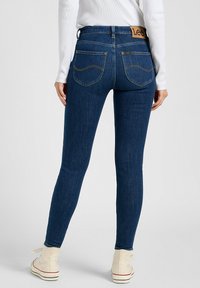 Jean en denim bleu foncé avec une taille mi-haute, des poches arrière et une patch en cuir brunavec le logo "Lee". Tissu doux et extensible ; design ajusté.