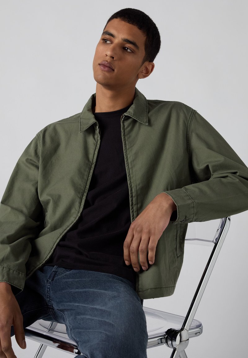 Joven sentado en una silla transparente, viste una chaqueta verde oliva, camisa negra y vaqueros azules, mirando ligeramente hacia arriba y a la izquierda.