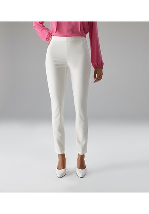 Frau trägt eng anliegende weiße Hose, weiße spitze Schuhe und eine rosa Langarmbluse, steht vor einem schlichten grauen Hintergrund.