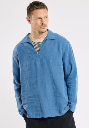 Homme aux cheveux bruns courts portant une chemise bleue à manches longues ample sur un sous-pull rayé beige et noir, les mains dans les poches, regardant vers la gauche.