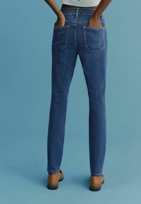 Jeans din denim de un albastru închis, cu un design drept, două buzunare la spate și o textură subtilă. Purtați cu pantofi slip-on de culoare maro.
