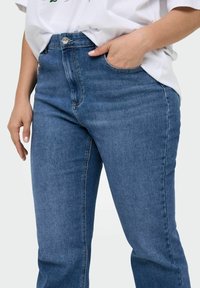 Jean en denim bleu taille haute avec une texture lisse, présentant un bouton frontale et des poches, visible au niveau de la taille et des cuisses.