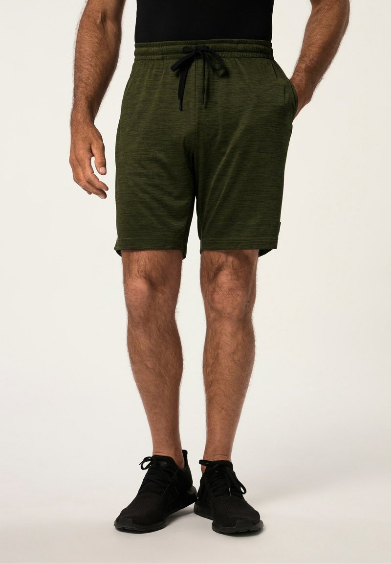 Homme portant un short vert foncé à cordon de serrage et des baskets noires, debout avec une main dans la poche sur un fond uni.
