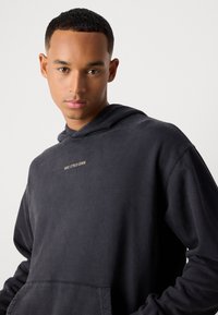 Ung mand med kort hår iført en mørkegrå Marc O'Polo Denim hoodie, der ser direkte fremad mod en ensartet lys baggrund.