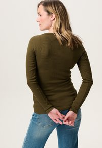 Olijfgroen lange mouwen ribbeltop met een ronde hals, getailleerde pasvorm en gestructureerde stof. Gepaard met blauwe jeans, handen achter de rug gekruist.