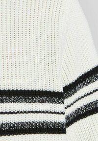 Weißer Strickpullover mit schwarzen horizontalen Streifen, der ein strukturiertes Muster aufweist. Das Garn ist leicht und atmungsaktiv, ideal zum Übereinandertragen.