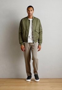 Olive green bomber jacket with front zip and pockets, worn over a white t-shirt, paired with light beige trousers and black sneakers.