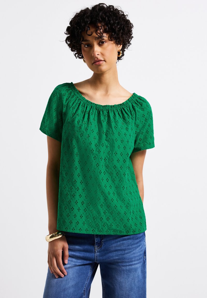 Top verde a maniche corte con scollatura arricciata, caratterizzato da un motivo in broderie anglaise. Realizzato in tessuto leggero, abbinato a pantaloni in denim.