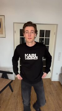 Giovane uomo che sistema una felpa nera Karl Lagerfeld, in piedi in una stanza con pavimento in legno e porte in vetro con cornice bianca.