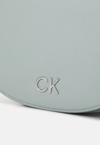 Světle modrá kožená taška přes rameno s texturovaným povrchem, s stříbrným logem "CK" a detailem prošití. Kulatý tvar s elegantním designem.