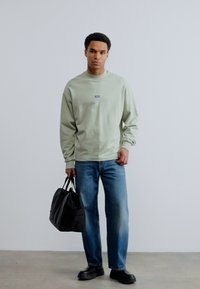 Άνδρας που φοράει ανοιχτό πράσινο φούτερ και μπλε τζιν, κρατώντας μια μαύρη τσάντα duffel, στέκεται μπροστά σε έναν απλό λευκό τοίχο.