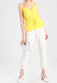 Top peplum amarillo sin mangas con escote en V, cintura cinchada, combinado con pantalones vaqueros cortos blancos y zapatos de tacón altos blancos, sosteniendo un clutch plateado.