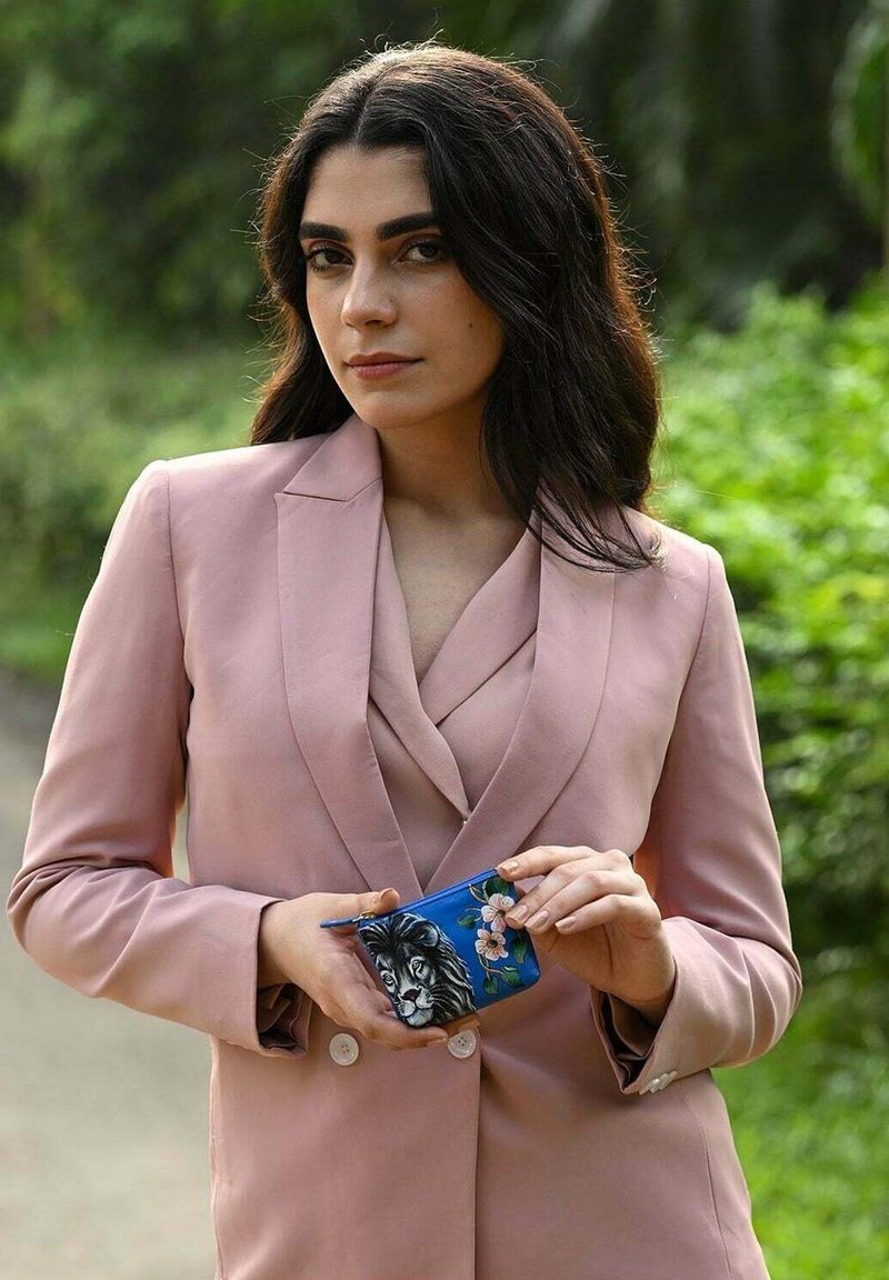 Blush roze op maat gemaakt pak met een reverskraag; vasthoudend een blauwe tas met een leeuw en bloemdesign. Achtergrond is groene bladeren.