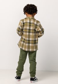 Grünes karierter Flanellhemd mit gelben und weißen Akzenten, kombiniert mit grünen Sweatpants und schwarzen Turnschuhen, von hinten gesehen.