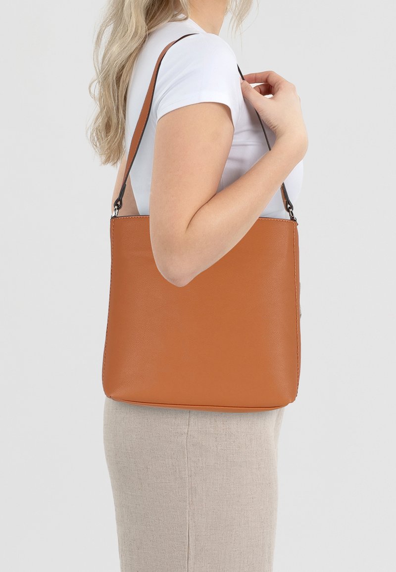 Sac à épaule en cuir tan avec une texture lisse, de forme rectangulaire et une sangle noire, porté par une personne en chemise blanche.