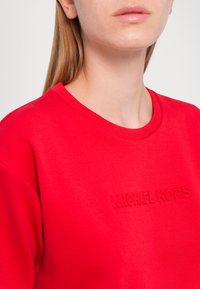 Sweatshirt roșu din bumbac, cu guler rotund, având un logo „MICHAEL KORS” embossat pe piept. Textură netedă, croială casuală.