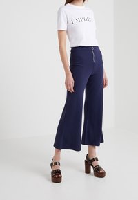 Witte t-shirt met korte mouwen en zwarte tekst, gecombineerd met marineblauwe wijde broek met een rits aan de voorkant, en bruine plateausandalen met gouden accenten.
