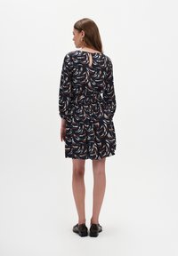 Kobieta stojąca tyłem, ubrana w granatową sukienkę z białymi i rdzawymi abstrakcyjnymi wzorami, do której założyła brązowe mokasyny i złote kolczyki.