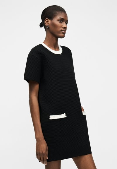 Frau mit dunkler Haut trägt ein kurzärmeliges, schwarzes, strukturiertes Kleid mit weißem Besatz an Kragen und Taschendetails, stehend vor einem einfarbigen Hintergrund.