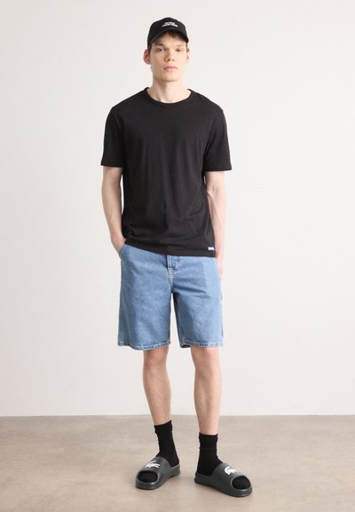 T-shirt noir à manches courtes, short en denim bleu, chaussettes noires et tongs noires avec des accents blancs. Le modèle se tient contre un fond uni.