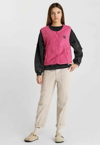 Roze fleecevest met een ronde halslijn en twee voorzakken, gedragen over een zwarte trui. Gecombineerd met beige ribfluwelen broek en crème schoenen.