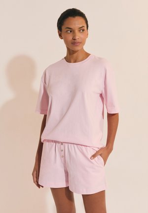 Femme portant un t-shirt rose clair à manches courtes et un short assorti, debout avec une main dans la poche, sur un fond beige clair.