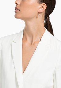 Blazer bianco con scollatura a V profonda, caratterizzato da rever strutturati e una superficie liscia. Orecchini a cerchio dorati con un piccolo accento blu.