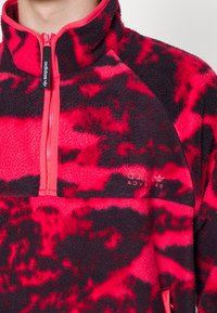 Pullover in pile rosso e nero con collo a zip, colletto a contrasto e logo "adidas Adventure". Presenta una superficie morbida e testurizzata e un motivo audace.