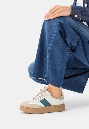 Persoon draagt witte en beige sneakers met groene strepen, blauwe jeans met wijde pijpen en een spijkerjack, zittend met handen rustend op de knie.