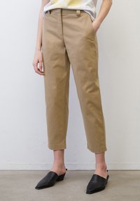 Osoba v béžových zkrácených kalhotách s bočními kapsami a černých špičatých nazouvacích botách, stojící u bílé zdi.